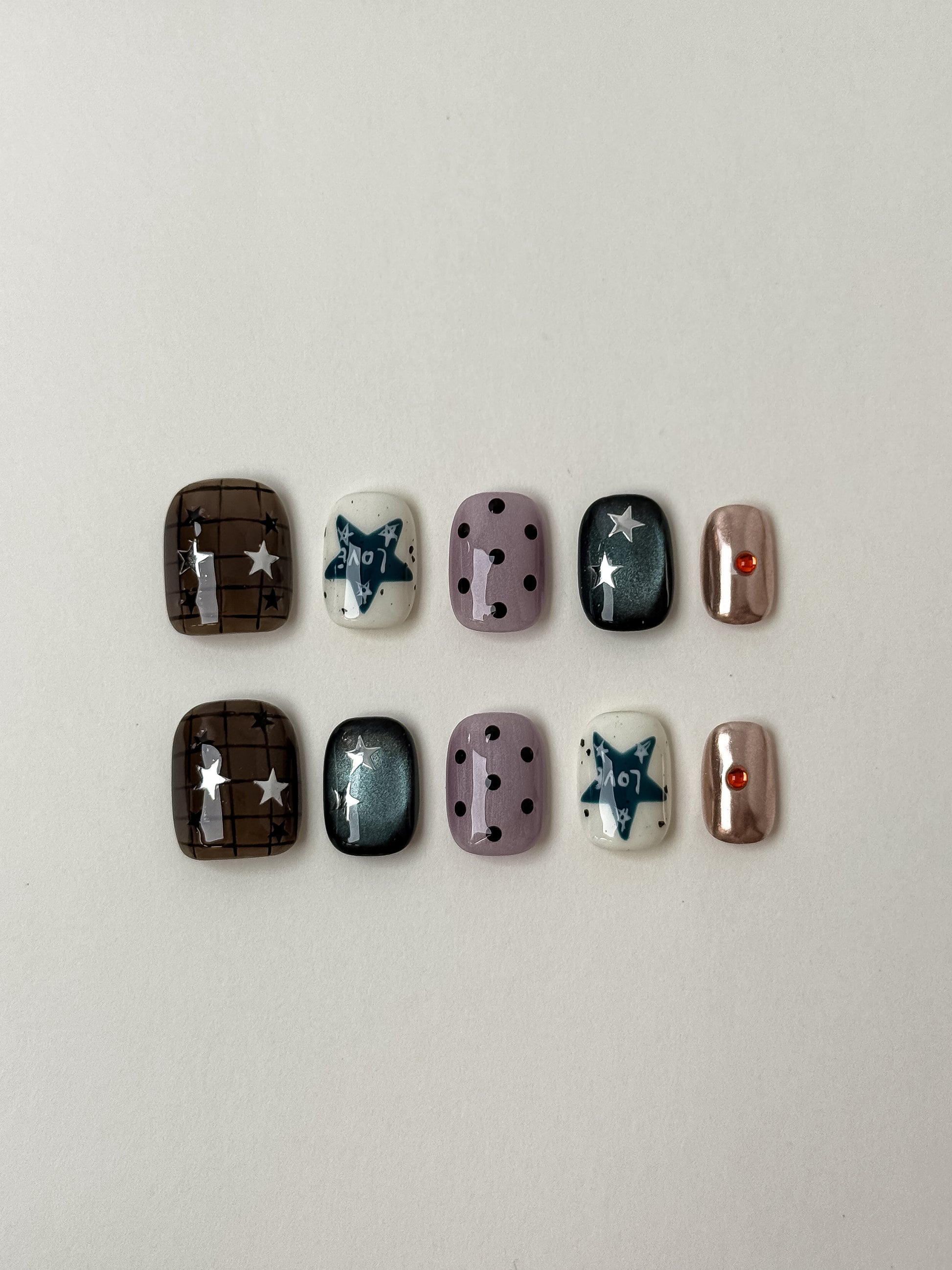 A complete set of ten press-on nails displayed on a white background featuring a mix of Y2K-inspired designs, including brown plaid, purple polka dots, teal cat-eye stars, and metallic rose gold with rhinestones.
